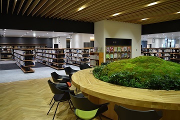 奥には背丈ほどの書棚が並んでおり、手前には大きな丸い机の周りに椅子が配置された図書館内部の写真