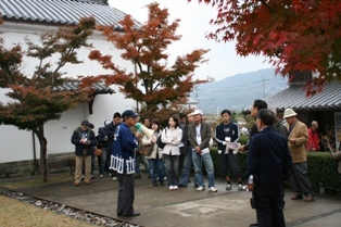 紅葉の木に囲まれた庭でボランティアガイドの話を聞く観光客