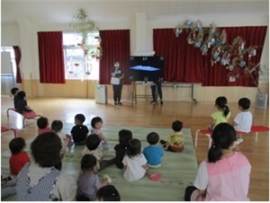 七夕の笹の葉が飾られた遊戯室で先生の話を聞く子供たち