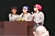割烹着を着た三人の女子高校生が舞台で話をしている写真。